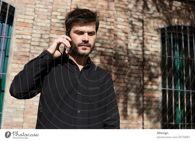 Young bearded man in sunglasses and denim shirt stands on city street and talks on his cell phone. casual attire lifestyle 1 male caucasian adult mobile young