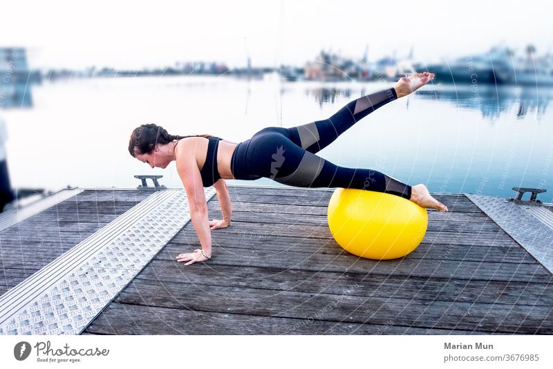 CHICA CON UNA PELOTA DE PILATES HACIENDO ESTIRAMIENTOS AMARILLA ESTIRAR DEPORTE AIRELIBRE PUERTO BELLEZA PIES AGUA DEPORTISTA JUVENTUD