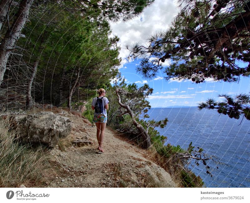 Young active feamle tourist wearing small backpack walking on coastal path among pine trees looking for remote cove to swim alone in peace on seaside in Croatia. Travel and adventure concept.