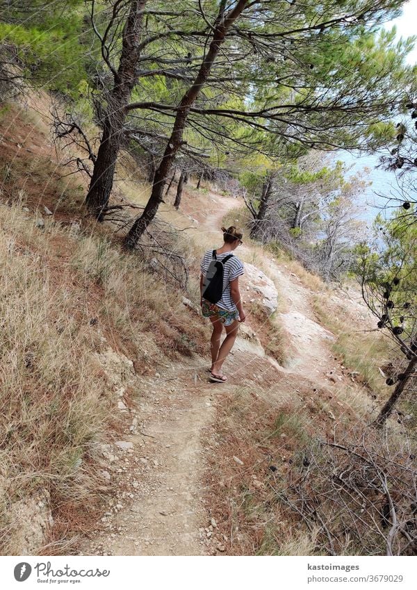 Young active feamle tourist wearing small backpack walking on coastal path among pine trees looking for remote cove to swim alone in peace on seaside in Croatia. Travel and adventure concept.