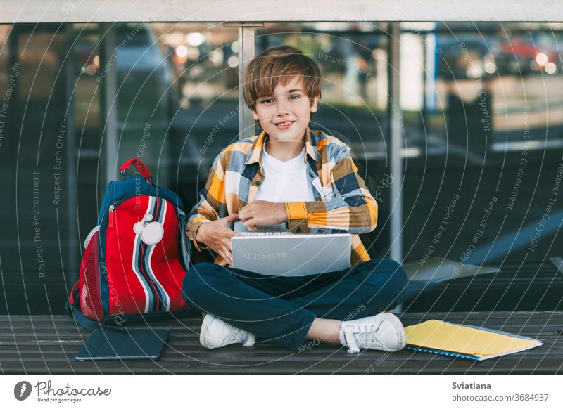 A cute boy in a plaid shirt is sitting on a bench with a laptop and typing on the keyboard, next to a backpack. The student is preparing for outdoor classes