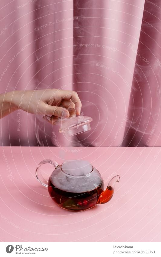 Female hand holding tea pot lid above kettle with the steaming fruit and berry tea beverage hot table hygge drink breakfast serving glass cover tranquillity