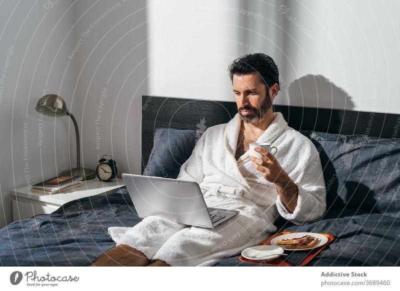 Calm man having breakfast on bed freelance coffee laptop work remote bathrobe using male businessman cup fresh morning drink device internet comfort online