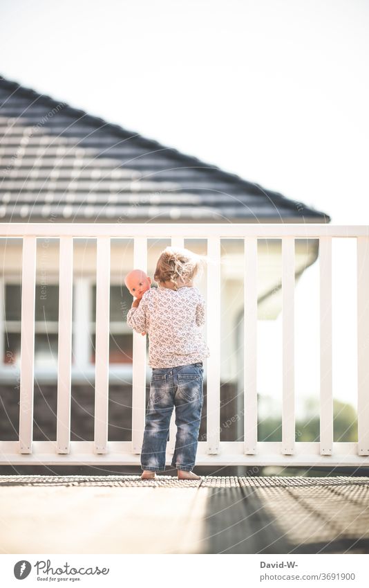 Girl with doll stands alone on the balcony and looks down from above Balcony girl peep out Grating Fence penned by oneself Doll Playing Observe Lonely being out