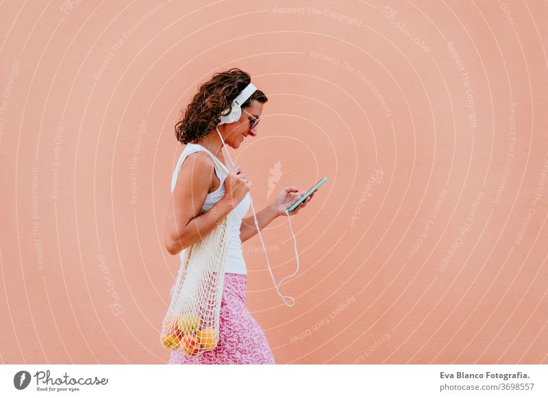 woman using mobile phone, listening to music on headset. walking by the city with cotton bag with fruit. Eco friendly, zero waste concept eco friendly shopping