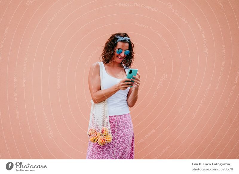 woman walking by the city, using mobile phone and holding a cotton bag with fruit. Eco friendly, zero waste concept eco friendly shopping technology caucasian