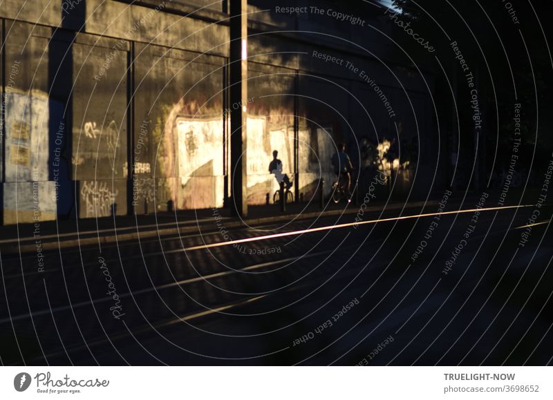 A cyclist disappears into the darkness next to the flashing railway tracks, but the low evening sun casts its sporty shadow on the concrete wall illuminated by the last rays of the sun