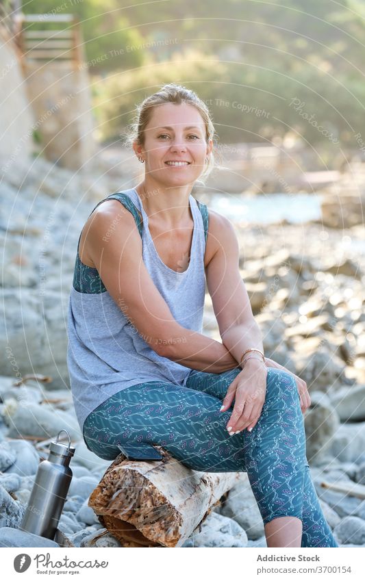 Carefree woman on stony beach in morning rocky seashore relax yoga chill smile sportswear female tree trunk sit rest happy harmony coast ocean wellbeing lady