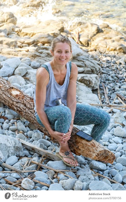 Carefree woman on stony beach in morning rocky seashore relax yoga chill smile sportswear female tree trunk sit rest happy harmony coast ocean wellbeing lady