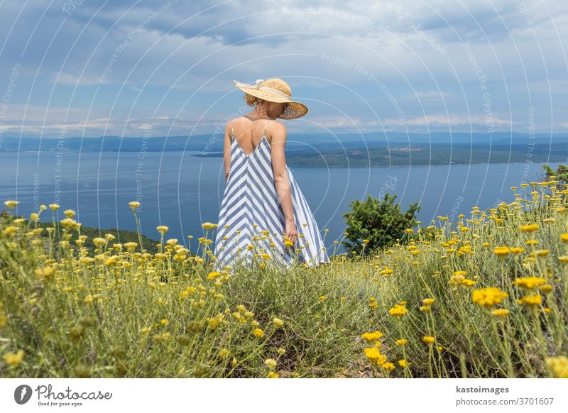 Rear view of young woman wearing striped summer dress and straw hat standing in super bloom of wildflowers, relaxing while enjoing beautiful view of Adriatic sea nature, Croatia