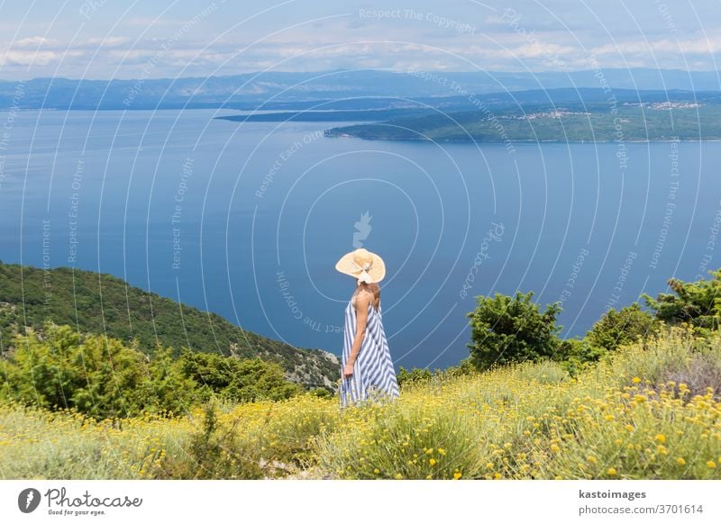 Rear view of a young woman in a striped summer dress and straw hat, standing in a super blossom of wild flowers and relaxing while enjoying a beautiful view of the Adriatic nature, Croatia