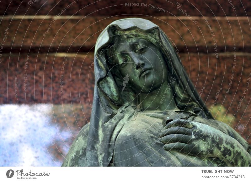 Blue-green shimmering sculpture of a beautiful woman in mourning, perhaps from the Art Nouveau era, in front of reddish-brown polished marble, in which a little blue sky with white clouds is reflected, but this does not harm the mourning