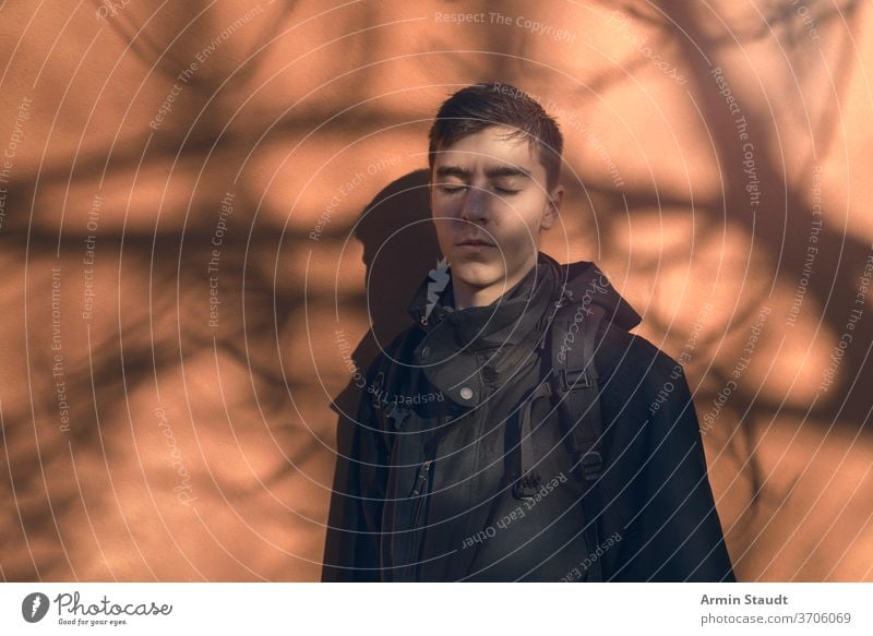 Portrait of a young man standing with his eyes closed in front of an orange wall with the shadow of a tree. sunbath sunlight outdoors relax leisure hands silent