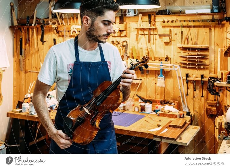 Male master in workshop with violins man repairman instrument music craftsman workbench apron male shiny equipment modern hobby tool focus table professional