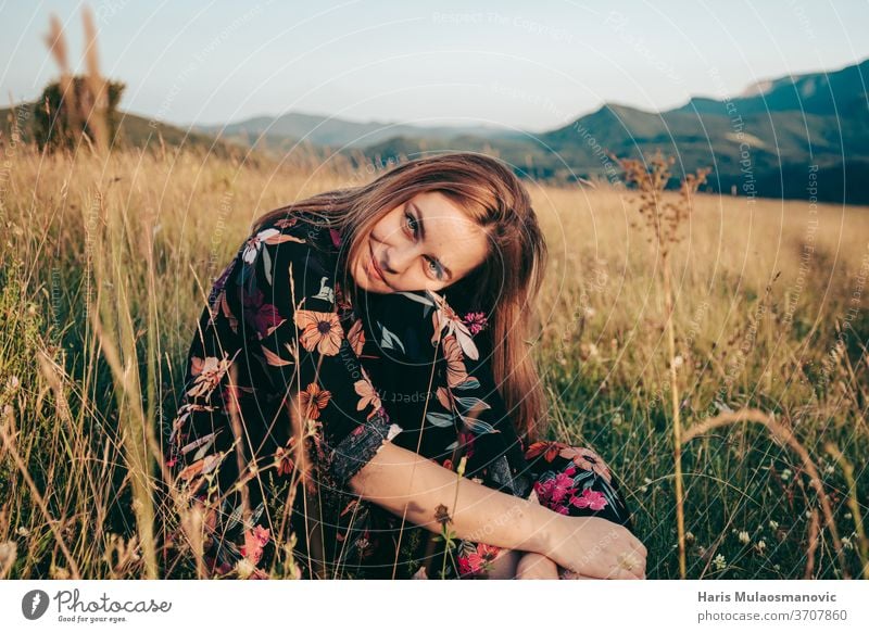 Happy woman sitting in grass field with mountains in background in sunset adventure beautiful beauty brown caucasian cute dress female flower freedom girl