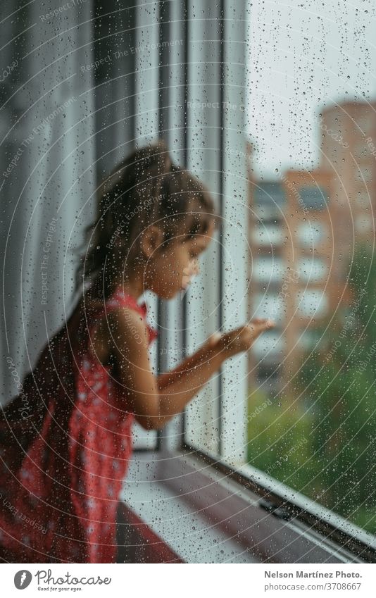 Little girl looking through the window in her bedroom in a rainy day. She is standing in a white chair, backward. We can see drops in the crystal. red child