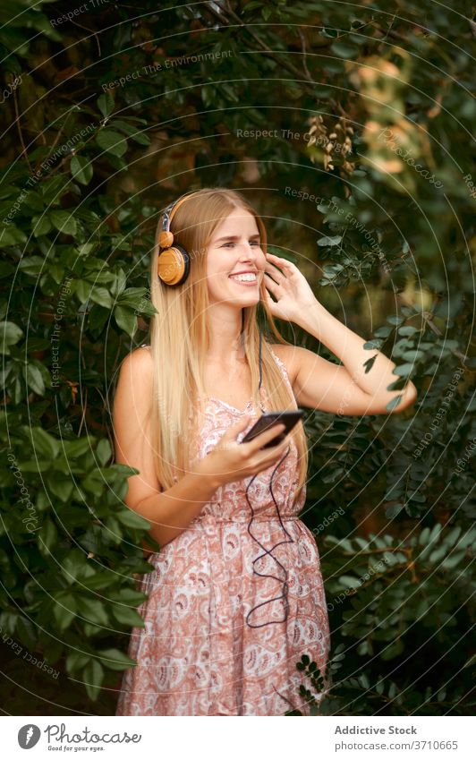 Happy young woman listening to music in garden smartphone headphones cheerful laugh having fun enjoy blond female summer foliage happy gadget device lifestyle