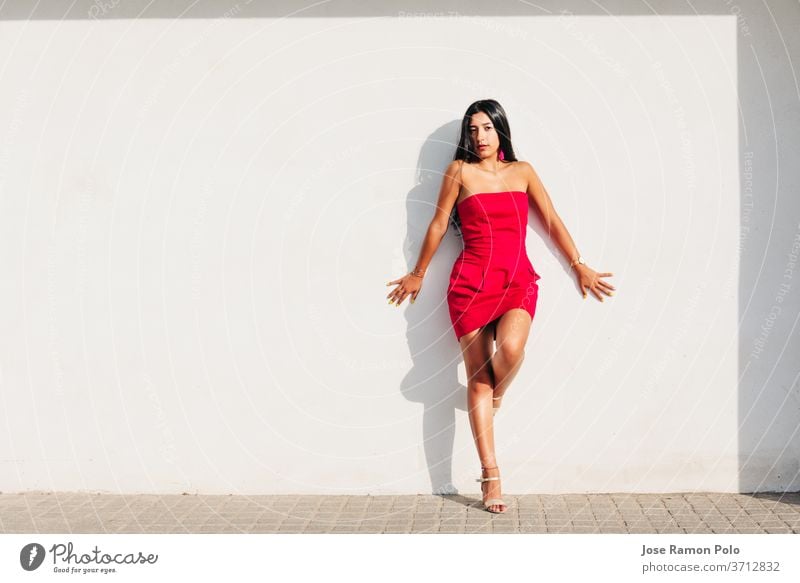 portrait of elegant latin young girl with long brunette hair and red dress on white wall with strong shadow, looking at camera White background beauty