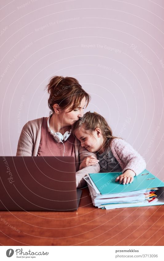 Woman working talking doing her job remotely during video chat phone call on laptop from home. Woman sitting at desk in front of computer looking at screen using headphones and smartphone while her daughter playing around. Concept of remote working