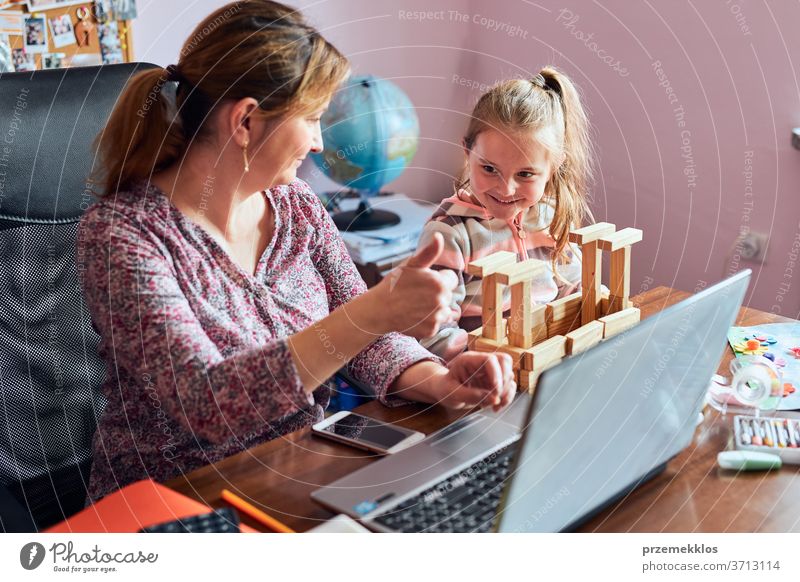 Woman mother working doing her job remotely during video chat call stream online course webinar on laptop from home while her daughter playing with bricks toy. Woman sitting at desk in front of computer looking at screen