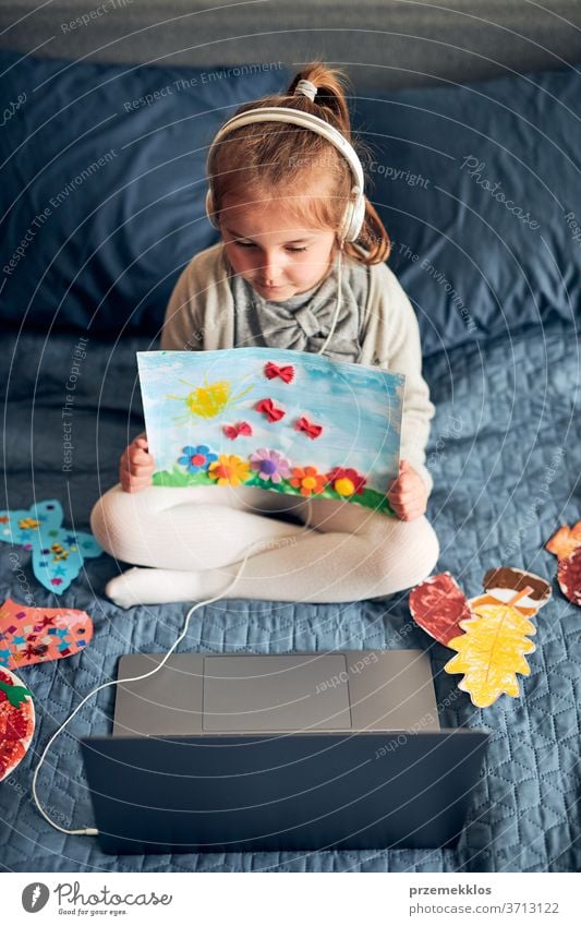 Little girl preschooler learning online showing her works drawings done at home. Child learning watching lesson remotely, talking with tutoress on video call from home during quarantine. Kid using laptop, headphones sitting on bed