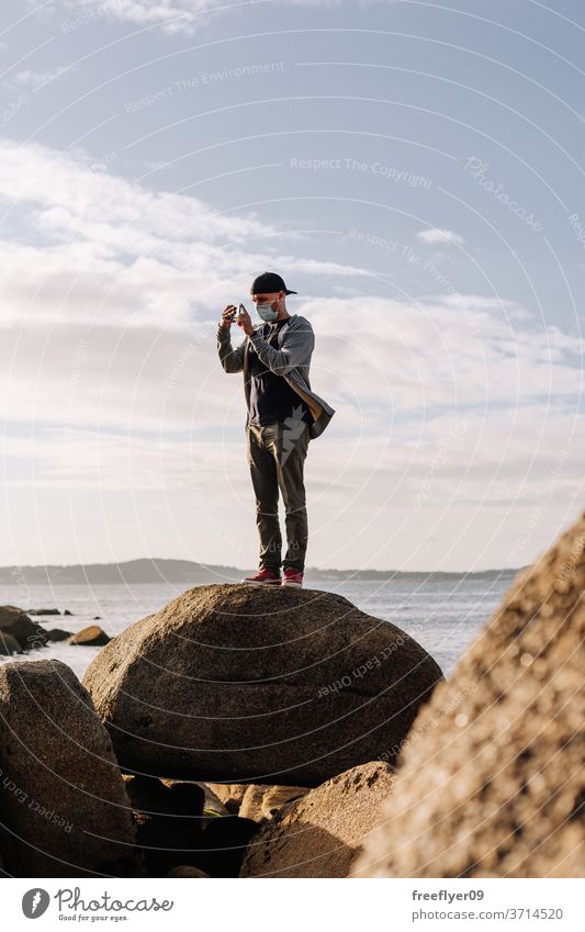 Tourist with a face mask taking photos with his smartphone tourist coast rock beach ocean portrait man surgical mask travel tourism traveler pandemic