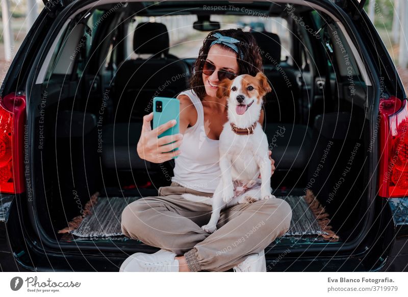 young happy woman in a car enjoying with her cute dog. Taking a selfie with mobile phone. Travel concept technology travel jack russell together love outdoors