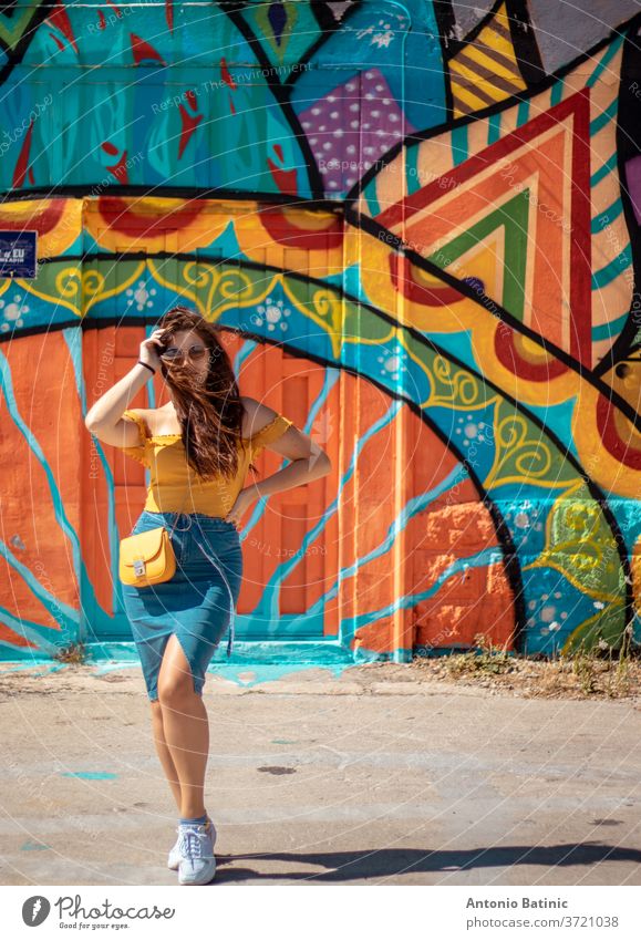 Attractive brunette girl wearing a blue skirt and an orange top posing on a summer day with a colorful wall in the background. Yellow purse, stylish influencer travelling and modelling