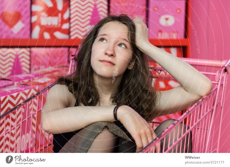 A young girl sits thoughtfully in a pink shopping trolley amidst pink goods Girl Shopping Trolley Consumption Consumer behaviour Pink gender roles Flashy