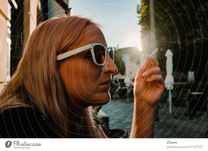 Young woman smoking in the sun Sun Summer Pedestrian precinct urban Town enjoyment Smoking Blonde long hairs Face Brown Cool sunglasses Lifestyle teen youthful