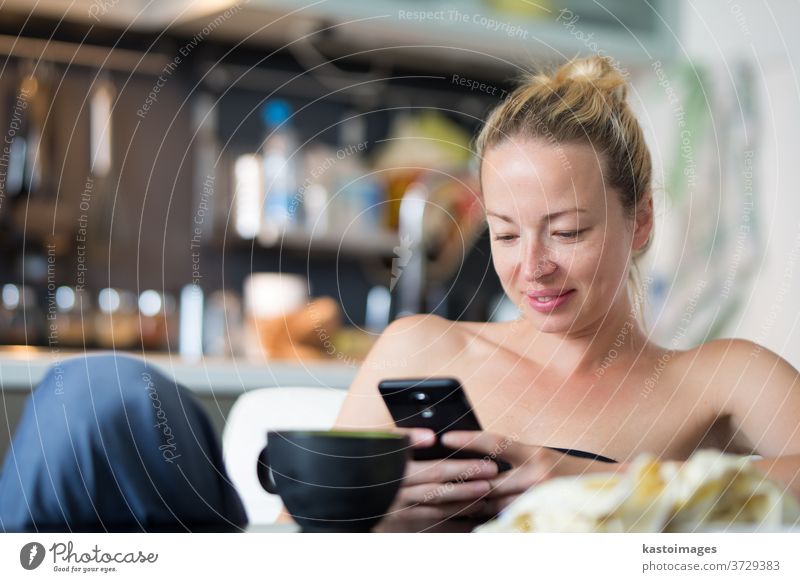 Young smiling cheerful pleased woman indoors at home kitchen using social media on mobile phone for chatting and staying connected with her loved ones. Stay at home, social distancing lifestyle.