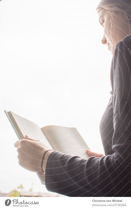 A young woman is sitting at an open window, holding an open book in her hands and reading it. Upper body and face in profile. Young woman Woman Profile