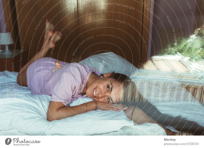 Portrait of pretty young girl looking at camera and laughing in morning and lying on white bed duvet, photographed through glass. sexy lip portrait woman