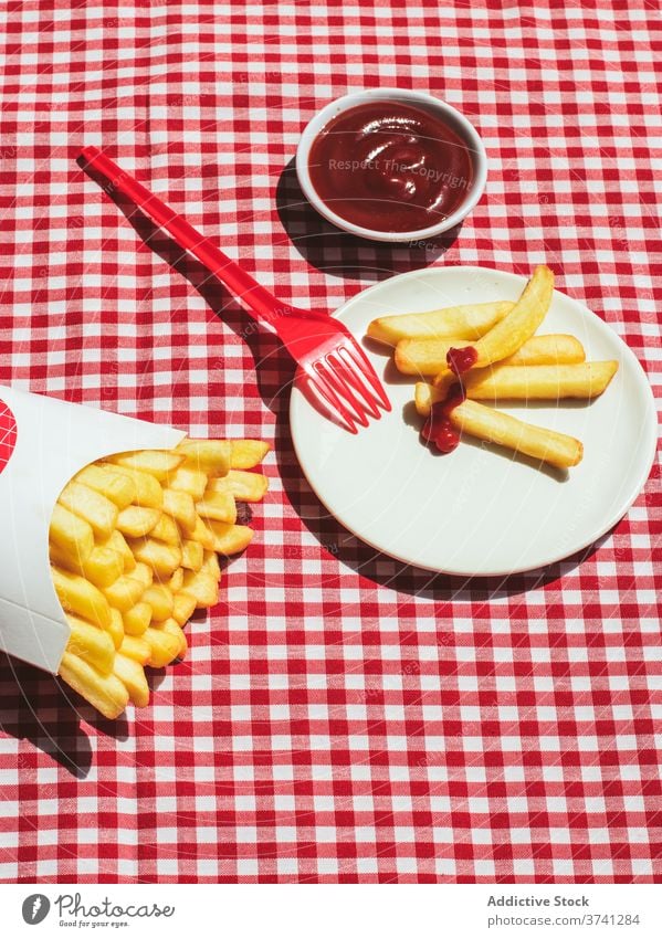 French fries packet near plate with potatoes soaked in ketchup american culture condiment potato chips unhealthy eating red background white frites take out