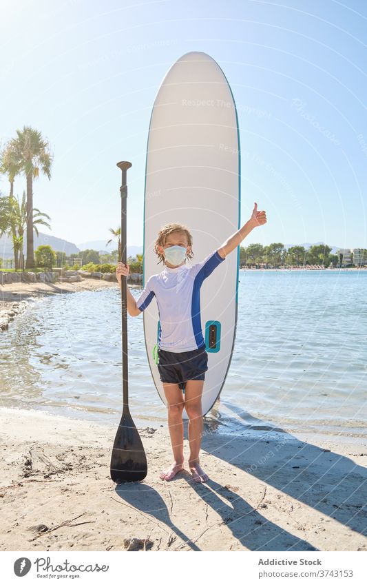 Child with a mask standing in front of a paddle surfboard while doing a gesture to be fine with the hand thunder breath pandemic prevention wear epidemic