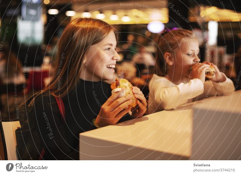 Beautiful schoolgirl eating hamburger sitting in a cafe indoors. Happy child eating junk food in a restaurant. Side view unhealthy fast food snack kid cute