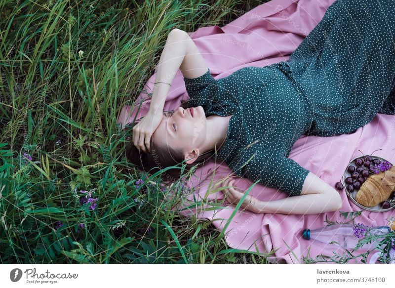 Portrait of young adult female in green dress lying on pink blanket with fruits, pastry and outdoors alone cherry croissant evening fabric flat lay food grass