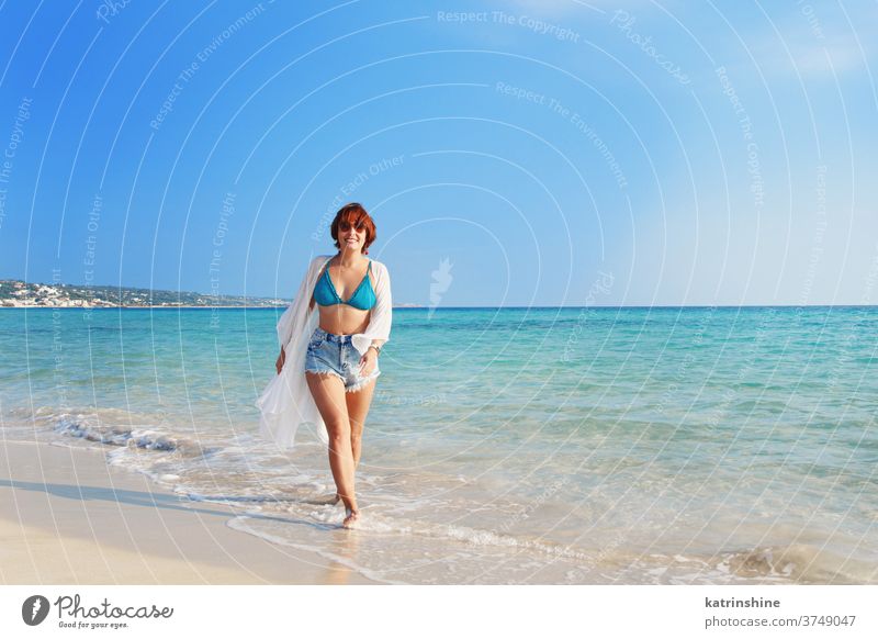 Young woman in cover up and jeans shorts walking on the beach kimono white blue turquoise highwaist Travel vacations concept destination outdoor happy enjoy