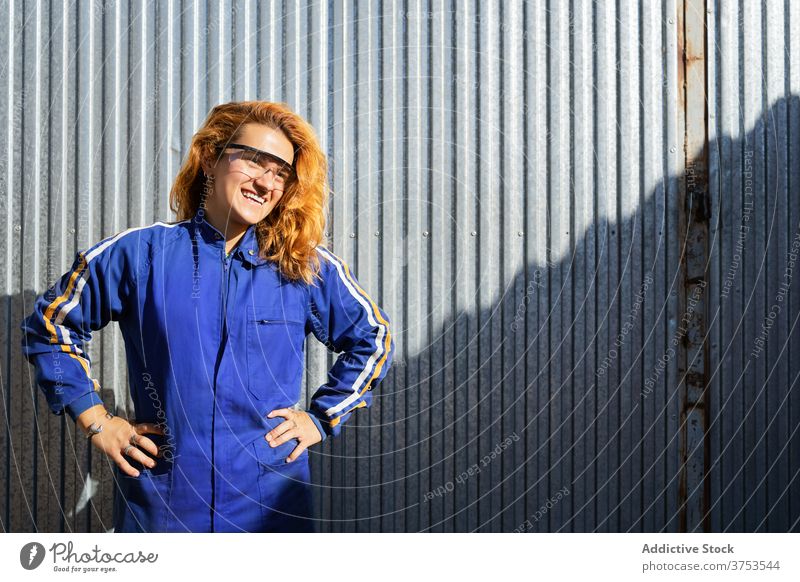 Content female worker in uniform near factory industrial woman employee break smile glasses plant workwear job metal wall professional joy rest confident lady