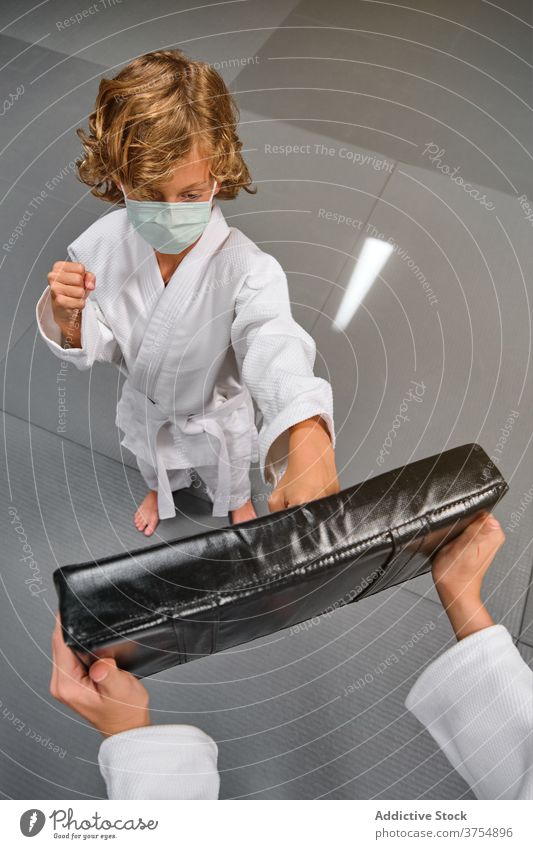 Boy in kimono and mask hitting a mitten with his fist in a gym impact sports skill trainer pose strength safe teamwork uniform wellness confident training