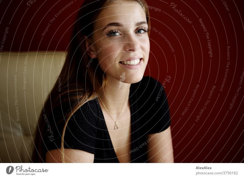 Young woman in a beige armchair in front of a red wall smiles Joy Day Aesthetics focused Force blue eyes décolleté great Definitive Calm athletic Skin