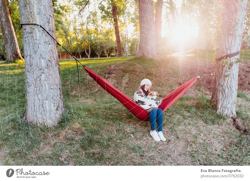 young woman relaxing with her dog in orange hammock. Covering with blanket. Camping outdoors. autumn season at sunset lying hammock jack russell pet nature park