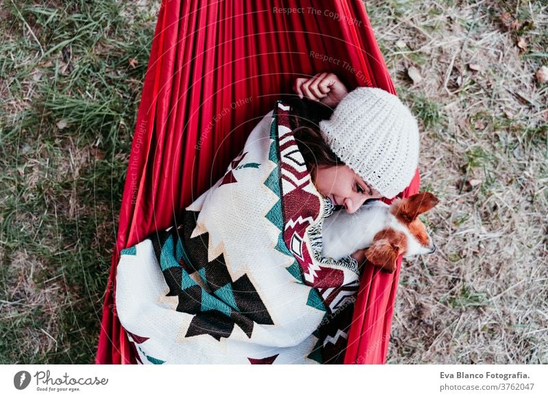 young woman relaxing with her dog in orange hammock. Covering with blanket. Camping outdoors. autumn season at sunset lying hammock jack russell pet nature park