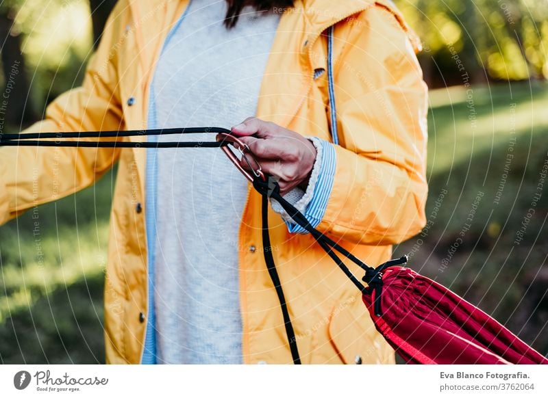 unrecognizable woman wearing yellow raincoat preparing hammock to relax. Camping outdoors. autumn season sunset orange park caucasian happy campground morning