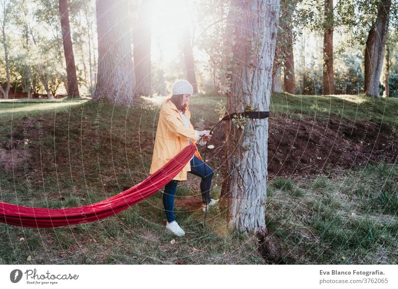 young woman wearing yellow raincoat preparing hammock to relax. Camping outdoors. autumn season sunset orange park caucasian happy campground morning light