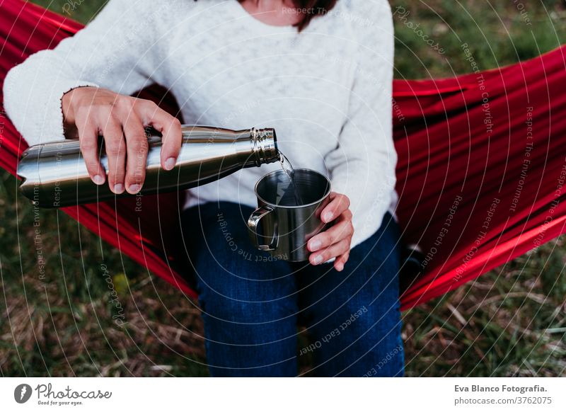 young woman holding thermos, pouring water into metallic mug, relaxing in hammock at sunset. autumn season unrecognizable feet lying outdoors nature orange park