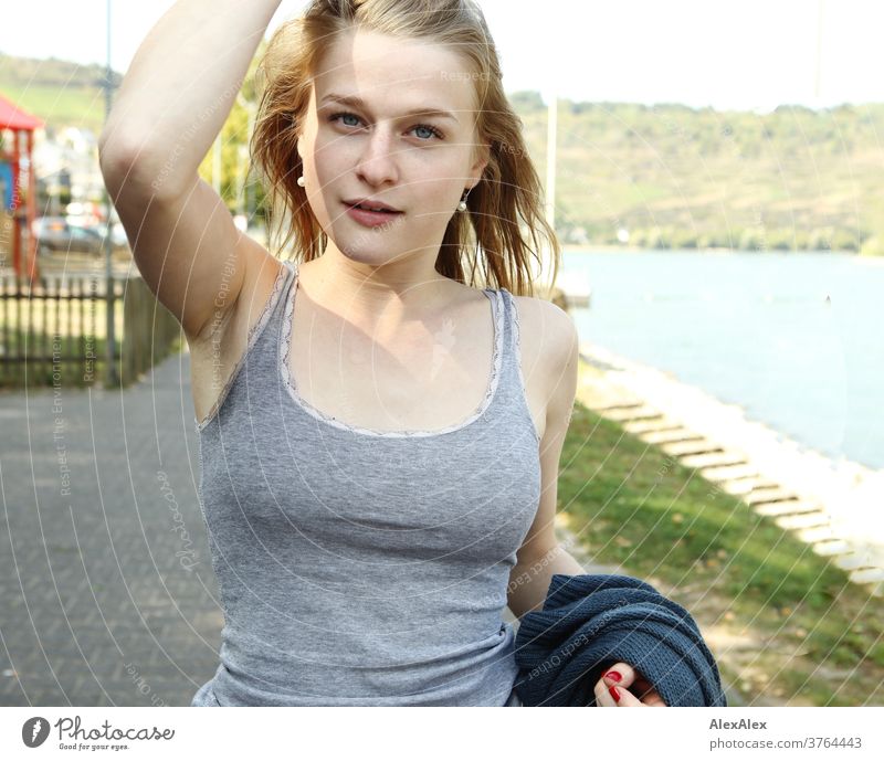 Young woman in a grey top touches her long blonde hair as she walks along the Rhine promenade Woman Slim already athletic Blonde youthful 18-25 years red blonde