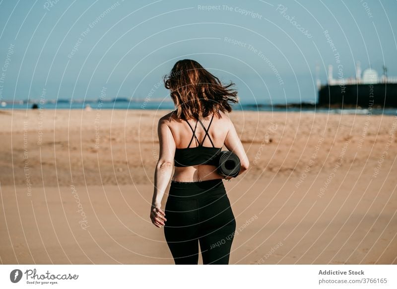 Anonymous woman with mat walking along beach yogi summer sportswear slim sunny yoga female water wellness harmony river riverside river bank shore activewear