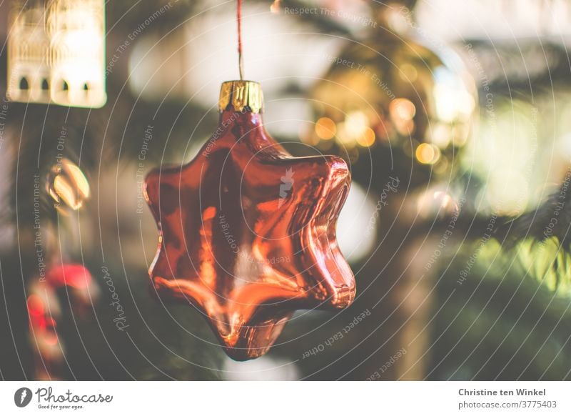 Christmas tree decorations made of glass in the shape of a star hang in the Christmas tree. In the background you see blurredly even more tree decorations