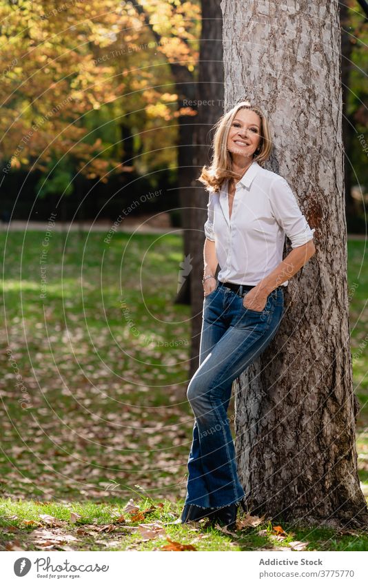 Smiling woman near tree in park stroll relax enjoy season sunlight garden carefree female autumn fall style recreation city casual rest trendy stand lady lean